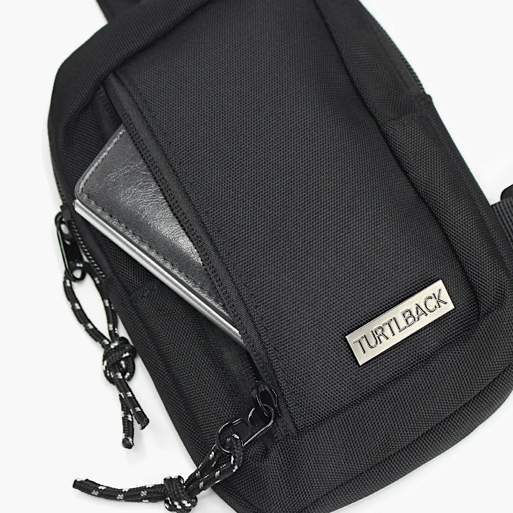 Geöffnete Vorderseite einer schwarzen mini Sling Bag mit Turtlback-Logo und herausragendem Portemonnaie, Kordel-Zipper-Verlängerung